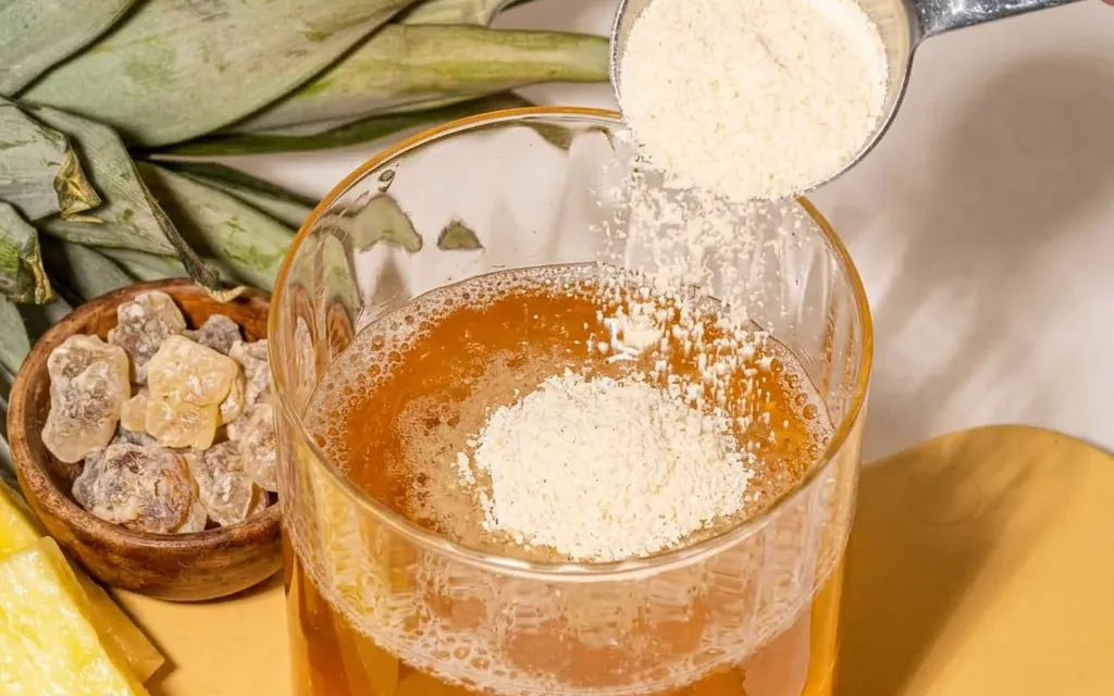 Cuillère doseuse versant une poudre blanche dans un verre de boisson ambrée.